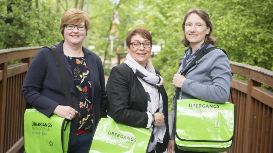3 Personen mit grünen Taschen auf denen ÜBergangsmanagement Schule - Beruf steht und das Logo der MaßArbeit zu sehen ist. 