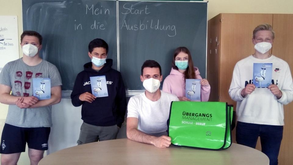 Klassenraum, 4 Schüler:innen vor Tafel, am Tisch sitzend Mann mit grüner Tasche Aufschrift "Übergangsmanagement Schule - Beruf"