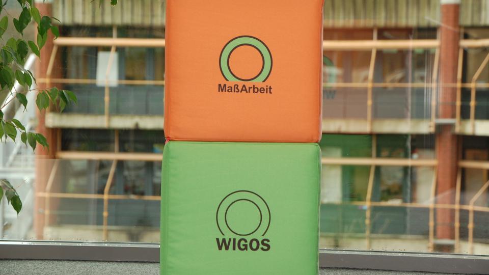 2 Würfel vor Kreishausfassadem Aufschrift oranger Würfel MaßArbeit, Aufschrift grüner Würfel Wigos
