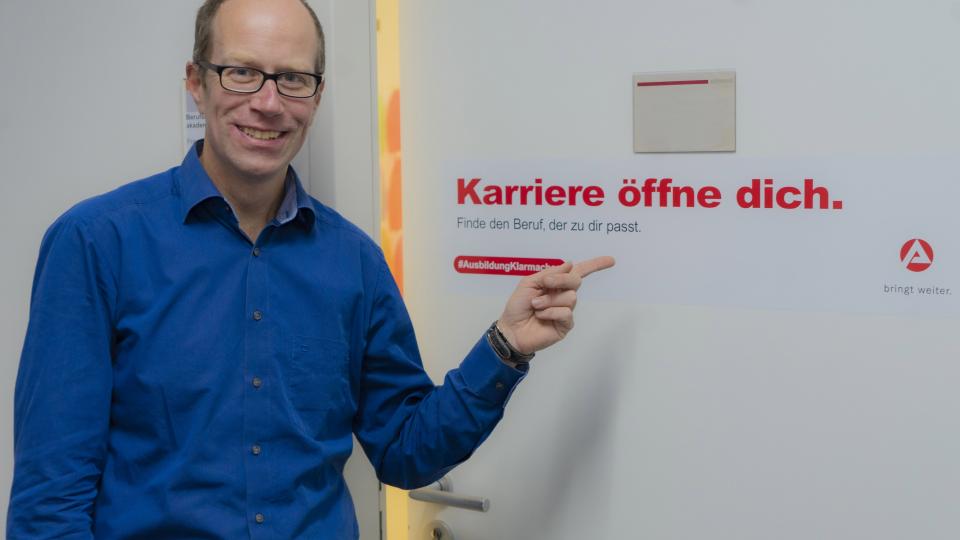 Herr Kampmeyer, Bereichsleiter der Berufsberatung der Agentur für Arbeit, zeigt mit dem Zeigefinger auf einen Aufkleber an der Tür mit dem Slogen "Karrierer öffne dich."