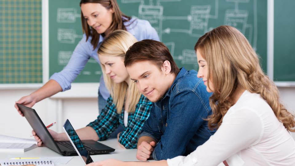 Mehrere Schüler sitzen an einem Tisch und schauen auf Laptops. Im Hintergrund ist eine Schultafel zu sehen.